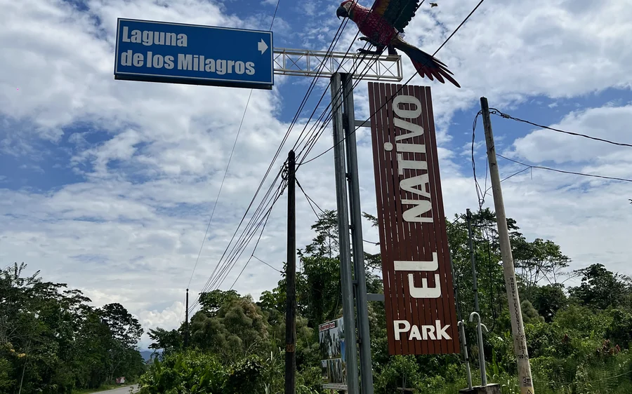 Letrero azul de la carretera que señala «Laguna de los Milagros» con una flecha, junto al cartel vertical de El Nativo Park coronado por un guacamayo